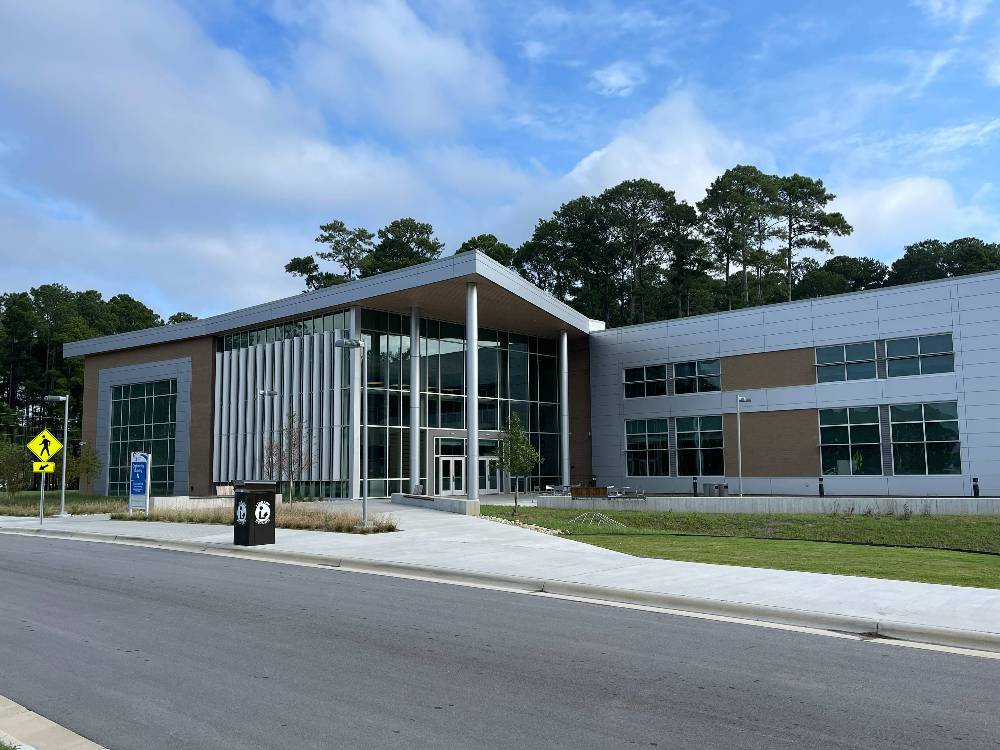 A modern two-story campus building with large glass windows and a covered entrance, surrounded by trees and a landscaped walkway under a partly cloudy sky.