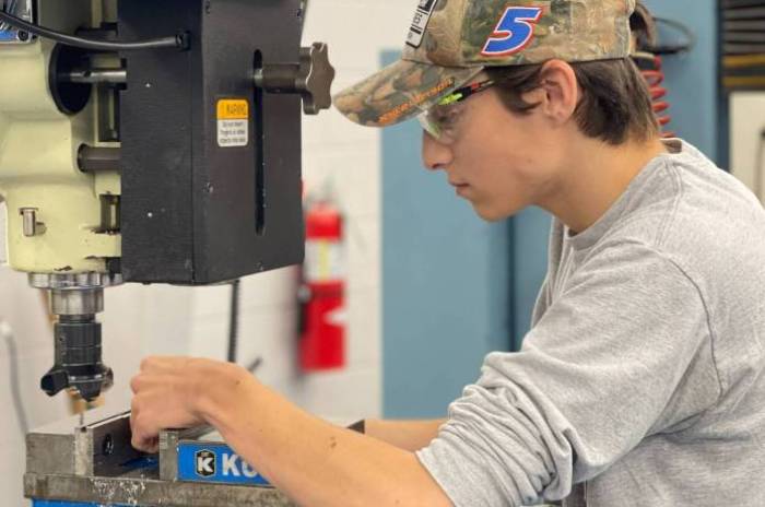 A student wearing safety glasses and a camouflage hat operates a milling machine in a workshop, using both hands to guide a metal piece secured in a blue vise.