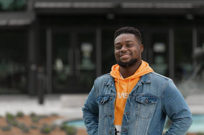 A student wearing an orange hoodie and denim jacket smiles while standing outdoors in front of a Tart campus building.
