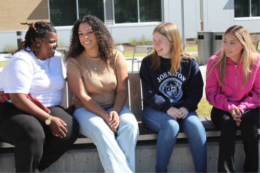 Four students sit together outdoors on a campus bench, talking and smiling in the sunlight.