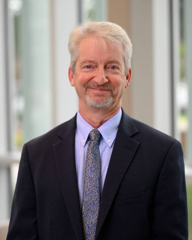 A professionally dressed man with light hair and a beard smiles while standing indoors in front of large windows.